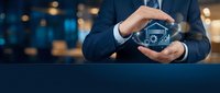 Professional businessman protecting house icon with hands in modern office environment representing real estate insurance and property protection services.