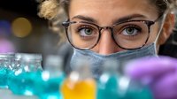 Young female scientist in protective mask and glasses examining colorful liquid samples in laboratory setting with blurred bokeh background.