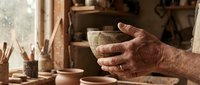 Skilled potter's weathered hands carefully holding handcrafted ceramic bowl in traditional pottery workshop with clay vessels and artisan tools.