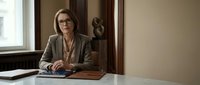 Professional middle-aged Caucasian businesswoman with glasses and beige blazer sitting at office desk, working on laptop in modern corporate workspace.