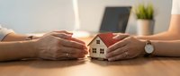 Two people protecting small wooden house model with hands on desk, symbolizing home insurance, real estate protection, and property security concept.