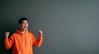 Man in orange hoodie celebrating success with fists raised against gray background, expressing joy and achievement for motivational and lifestyle content.