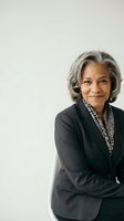 Confident mature African American businesswoman with gray hair sitting with arms crossed, wearing a blazer, smiling against light background.