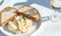 Creamy potato salad garnished with fresh herbs served on white plate with crusty bread slices and white wine glass, bright natural lighting creates soft shadows.