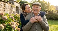 Happy mature couple embracing in garden during golden hour sunset, showing love and affection in outdoor setting with warm natural lighting and flowers.