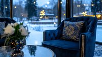 Luxurious navy blue velvet armchair with decorative pillow near window overlooking snowy winter landscape, cozy evening atmosphere with bokeh lights and floral decor.
