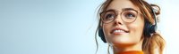 Young Asian woman in glasses and orange sweater wearing headphones smiles while listening to music against light blue background. Professional lifestyle portrait.
