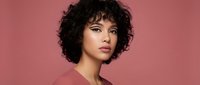 Young mixed-race woman with curly black hair and natural makeup posing confidently against pink background for beauty and fashion campaigns.