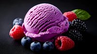 Vibrant scoop of berry ice cream surrounded by fresh blueberries, raspberries and blackberries with mint leaf on dark background, macro food photography.