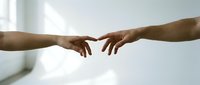 Two people with medium to dark skin tone reaching toward each other with fingertips nearly touching in a gesture of connection, support, or agreement against a bright, blurred background.