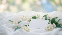 Blank white card surrounded by fresh jasmine flowers and green leaves on soft white fabric, perfect for wedding invitations, greeting cards, and spring event designs.