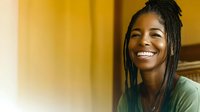 Young African American woman with braided hair and bright smile wearing casual green sweater against warm yellow background, radiating joy and confidence.