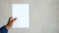 Man with dark skin tone holding blank white paper sheet against neutral gray wall background for mockup and branding purposes.