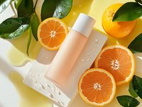 Cosmetic bottle with water drops surrounded by fresh cut oranges and green leaves on yellow background, natural skincare product photography styling.