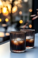 Two elegant ribbed glass tumblers filled with dark cocktails on table against blurred golden bokeh lights background, creating cozy evening atmosphere.