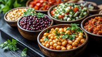 Variety of legumes and beans in wooden bowls with fresh herbs and vegetables, colorful healthy protein sources arranged on dark background for food photography.