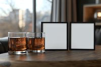 Two glasses of whiskey or bourbon on wooden table with empty black photo frames against window background, creating moody atmosphere for memories and reflection.