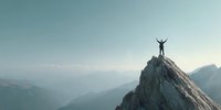 Silhouette of person with raised arms standing on mountain peak overlooking misty valley and distant ranges at dawn.