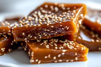 Homemade caramel candy pieces topped with sesame seeds on white plate, macro food photography showing rich golden brown color and sticky texture.