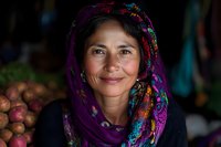 Middle-aged woman wearing colorful floral headscarf smiles warmly at camera, natural lighting highlights her genuine expression against market produce background.