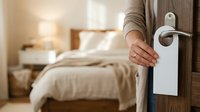 Person holding do not disturb sign on hotel room door with bedroom visible in background.