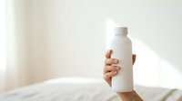 Unrecognizable person holding white cosmetic bottle with blank label in bright sunlit bedroom for skincare product marketing.