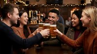 Group of diverse young adults toasting with beer glasses in cozy pub with string lights, celebrating friendship and social gathering.