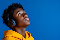 Young African American woman in yellow hoodie wearing headphones looking up with hopeful expression against vibrant blue background, modern lifestyle portrait.