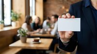 Businessman in dark suit holding blank white business card at office meeting with colleagues in background, networking and corporate communication concept.