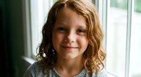 Happy young Caucasian girl with curly blonde hair smiling at camera by window. Child portrait showing joy and innocence in natural indoor lighting.