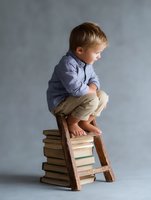 Caucasian toddler boy in blue shirt squatting on wooden ladder placed on stack of vintage books against gray background, barefoot studio portrait for education concept.
