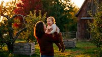 Mother lifting happy toddler boy in autumn farm setting with golden sunset light creating warm family moment outdoors in countryside.