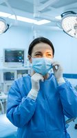Female surgeon putting on protective face mask in modern operating room with medical equipment and surgical lights preparing for procedure.