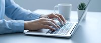 Professional businesswoman typing on modern laptop computer at clean white desk workspace with coffee cup and small plant in bright office environment.