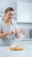 Female baker dusting flour on fresh braided bread loaf in modern bakery kitchen. Caucasian woman wearing apron preparing artisan baked goods for sale or commercial use.