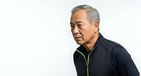 Senior Asian man with gray hair wearing black athletic jacket with green trim, looking downward in contemplative pose against white background.