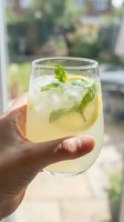 Hand holding glass of refreshing lemonade with fresh mint leaves and ice cubes outdoors on sunny day. Perfect for beverage, summer drink, and lifestyle content.