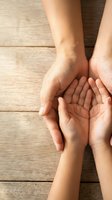 Adult hands gently protecting child hands in caring gesture on wooden background symbolizing family support, guidance and protection.