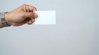 Male hand holding blank white business card or paper against neutral gray background for branding and corporate identity mockup purposes.