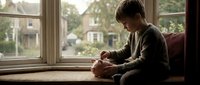Young child with light skin tone sitting on windowsill, holding teddy bear, wearing casual clothing, gazing outdoors through large window with suburban houses visible in background.