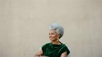 Smiling senior woman with gray hair wearing green velvet shirt against neutral beige wall background. Portrait of mature female for lifestyle and wellness content.