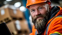 Mature bearded construction worker in orange safety vest and hard hat smiling while working at warehouse site, displaying confidence and expertise in industrial setting.