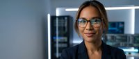 Professional Hispanic businesswoman with glasses smiling confidently in modern office environment with computer monitors and technology background.