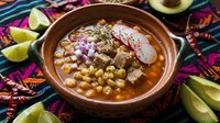 Traditional Mexican pozole soup with hominy corn, tender pork, radishes and lime in authentic clay bowl on colorful textile background for restaurant menus.