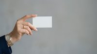 Mature man with medium skin tone holding blank white business card in hand against neutral gray background for corporate branding and marketing purposes.