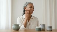 Mature African American woman applying face cream, smiling during skincare routine at home with ceramic jars on table.
