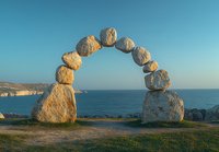Natural stone arch formation on coastal cliff edge with ocean view and blue sky at sunset, balanced rocks creating artistic gateway overlooking Mediterranean seascape.
