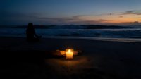 Peaceful evening beach scene with glowing candle in sand creating warm light circle while person sits silhouetted against twilight ocean waves and colorful sunset sky.