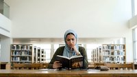 Young Middle Eastern man wearing a light blue hijab and dark cardigan reading an open book at a wooden library table with shelves in background, suitable for education, literacy, and learning concepts.