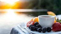 Fresh berries and hot coffee cup on white plate against blurred lake sunrise background, morning breakfast scene with natural sunlight and bokeh effect.